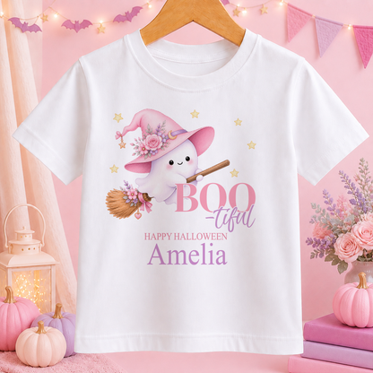 A white t-shirt with a cartoon ghost wearing a witch's hat and holding a broom, surrounded by pink and purple decorations, with the text "BOO TIFUL" and "HAPPY HALLOWEEN Amelia" printed on it.