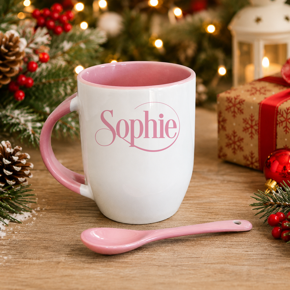 A white mug with a pink interior and handle, featuring the name "Sophie" in pink cursive text, sits on a wooden surface next to a pink spoon. The mug is surrounded by Christmas decorations, including pine cones, berries, and a lantern, creating a festive atmosphere.