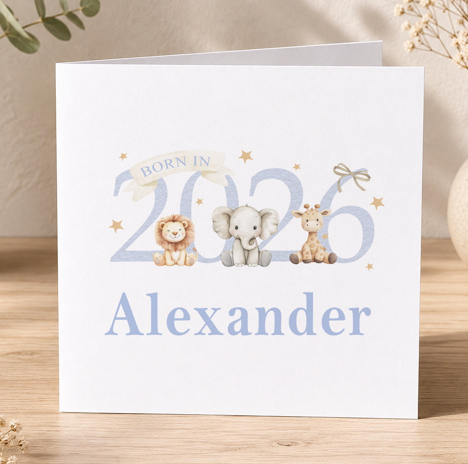 White square greeting card with a lion, elephant and giraffe cub sitting beside the number 2026, with the personalised name Alexander printed below, on a wooden surface with foliage.