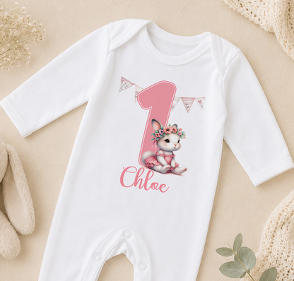 A white onesie with a pink number one and a small pink bunny wearing a flower crown, along with the name "Chloe" written on it.