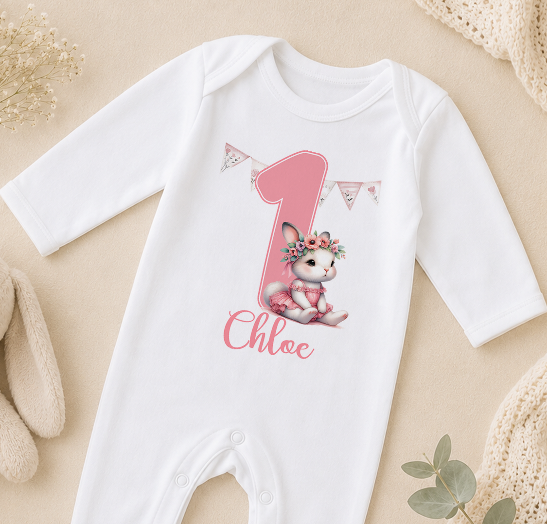 A white onesie with a pink number one and a small pink bunny wearing a flower crown, along with the name "Chloe" written on it.