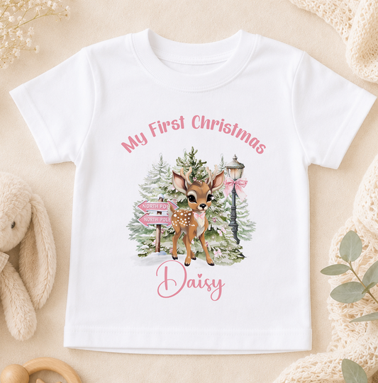 A white t-shirt with a printed design of a deer standing in front of a Christmas tree, with a sign that says "My First Christmas Daisy".