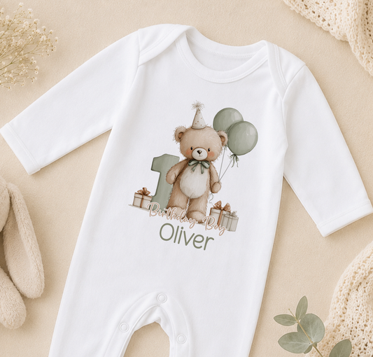 A white onesie with a teddy bear and balloons, along with the text "Olivier" printed on it, is displayed on a beige surface.
