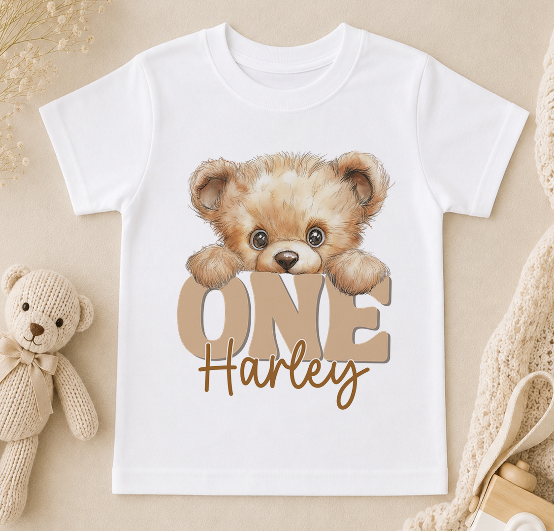 A white t-shirt with a teddy bear and the text "ONE Harley" printed on it, along with a small stuffed bear.