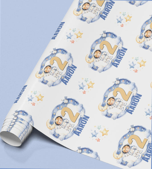 A roll of wrapping paper with a repeating pattern of a boy in a spacesuit, stars and clouds, shown here personalised with the age 2