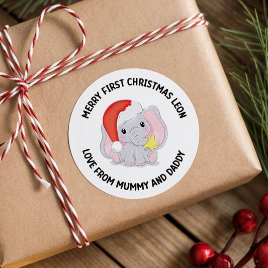 Round pastel Christmas sticker featuring a cartoon elephant wearing a red Santa hat surrounded by yellow stars and 'Merry First Christmas' wording, personalised with a custom name, on a brown baby gift box