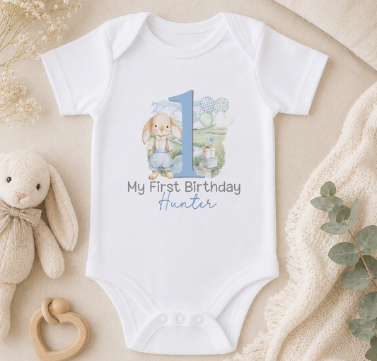 A white onesie with a blue bunny and the number 1, along with the text "My First Birthday Hunter" printed on it, is displayed on a bed surrounded by stuffed animals and a blanket.