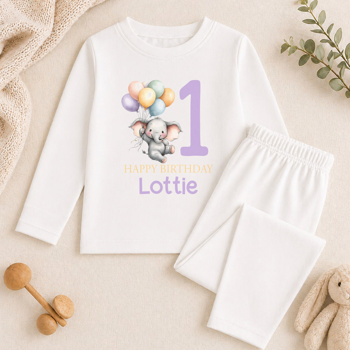 A white long-sleeved shirt with a graphic of an elephant and balloons, along with the text "Happy Birthday Lottie" printed on it. The shirt is placed on a beige surface, and there are some toys and a plant nearby.
