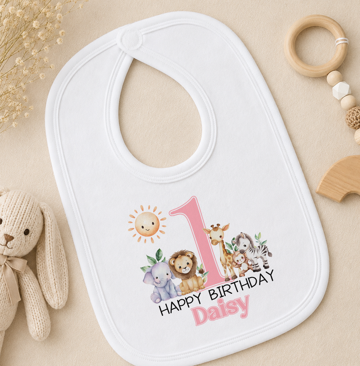 A white baby bib with a personalized print of a first birthday theme, featuring a sun and animals, is placed on a beige surface alongside a stuffed animal and baby toys.