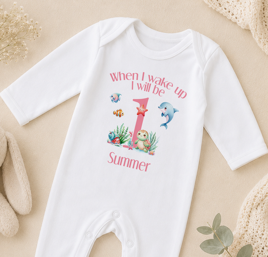 A white onesie with a printed design of a dolphin and other sea creatures, along with the text "When I wake up I will be 1 Summer".
