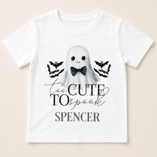 A white t-shirt with a graphic design featuring a ghost character wearing a bow tie, surrounded by bats, and the text "Too Cute to Spook" and "Spenser" below.