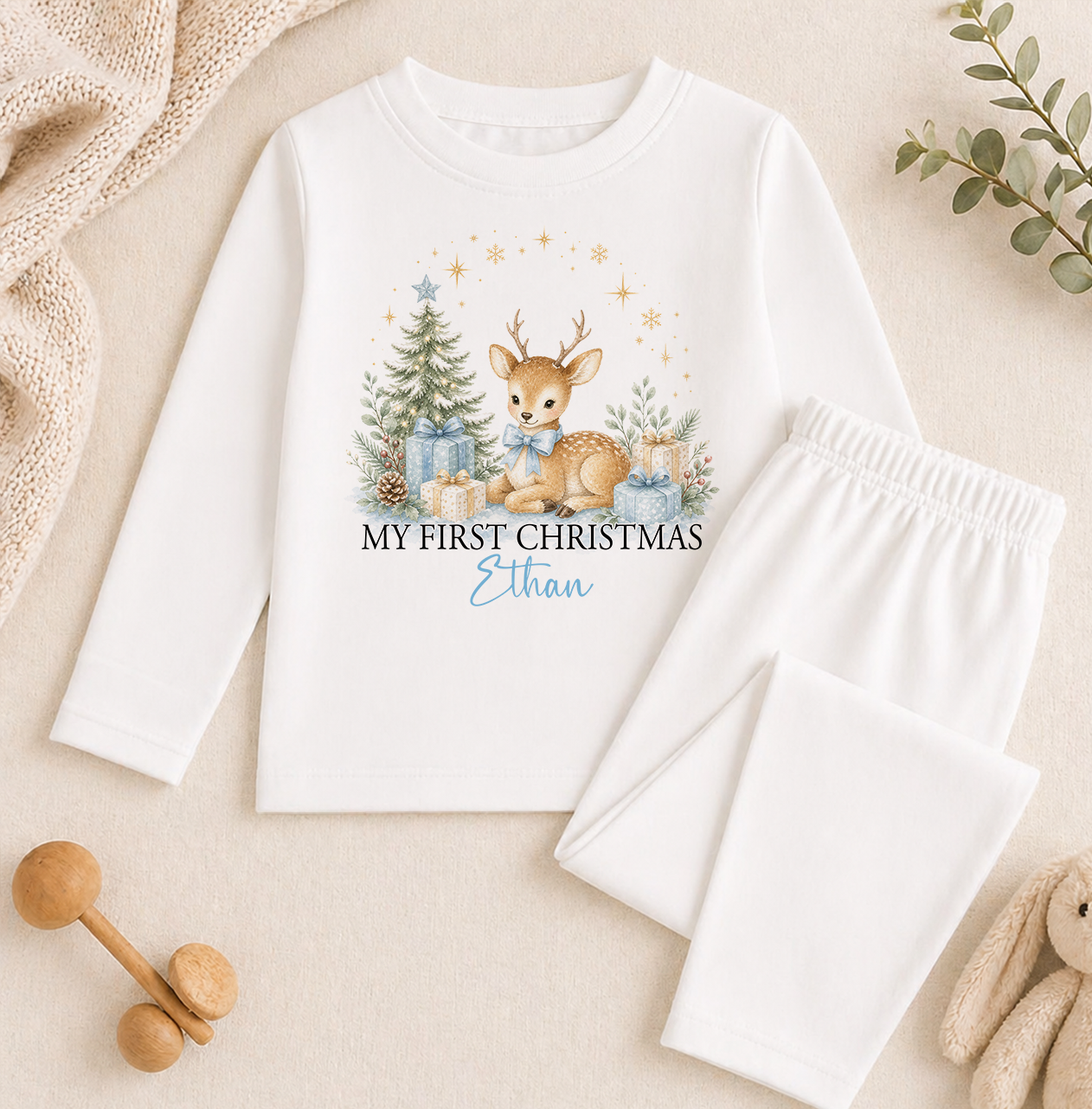 A white long-sleeved shirt with a Christmas-themed design, including a deer, presents, and stars, along with the text "My first Christmas Ethan". The shirt is placed on a beige background, and there are some toys and a plant nearby.