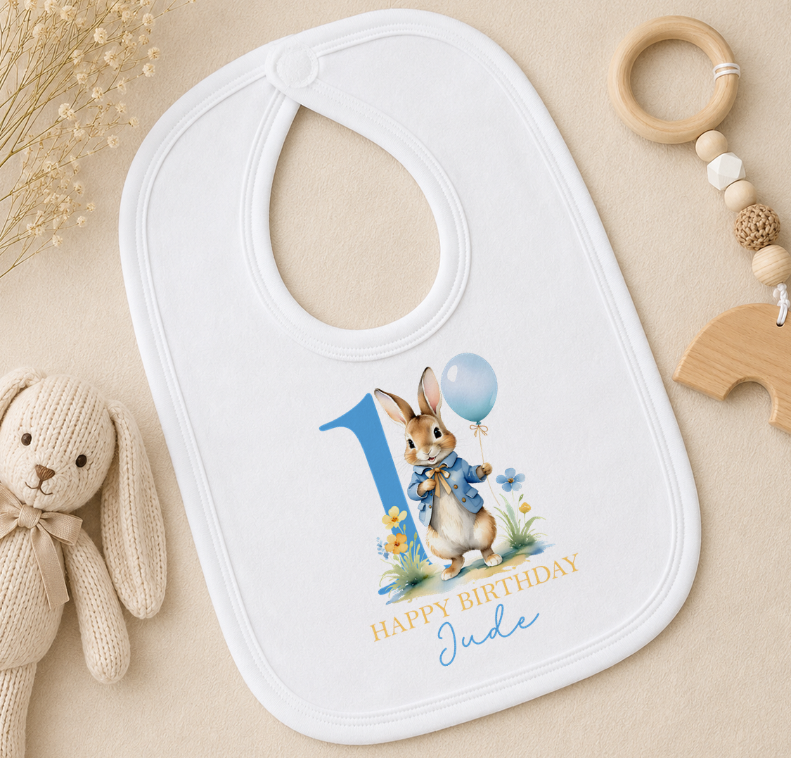 A white bib with a bunny wearing a blue outfit and holding a blue balloon, along with a wooden toy and a beaded rattle, all arranged on a beige surface.
