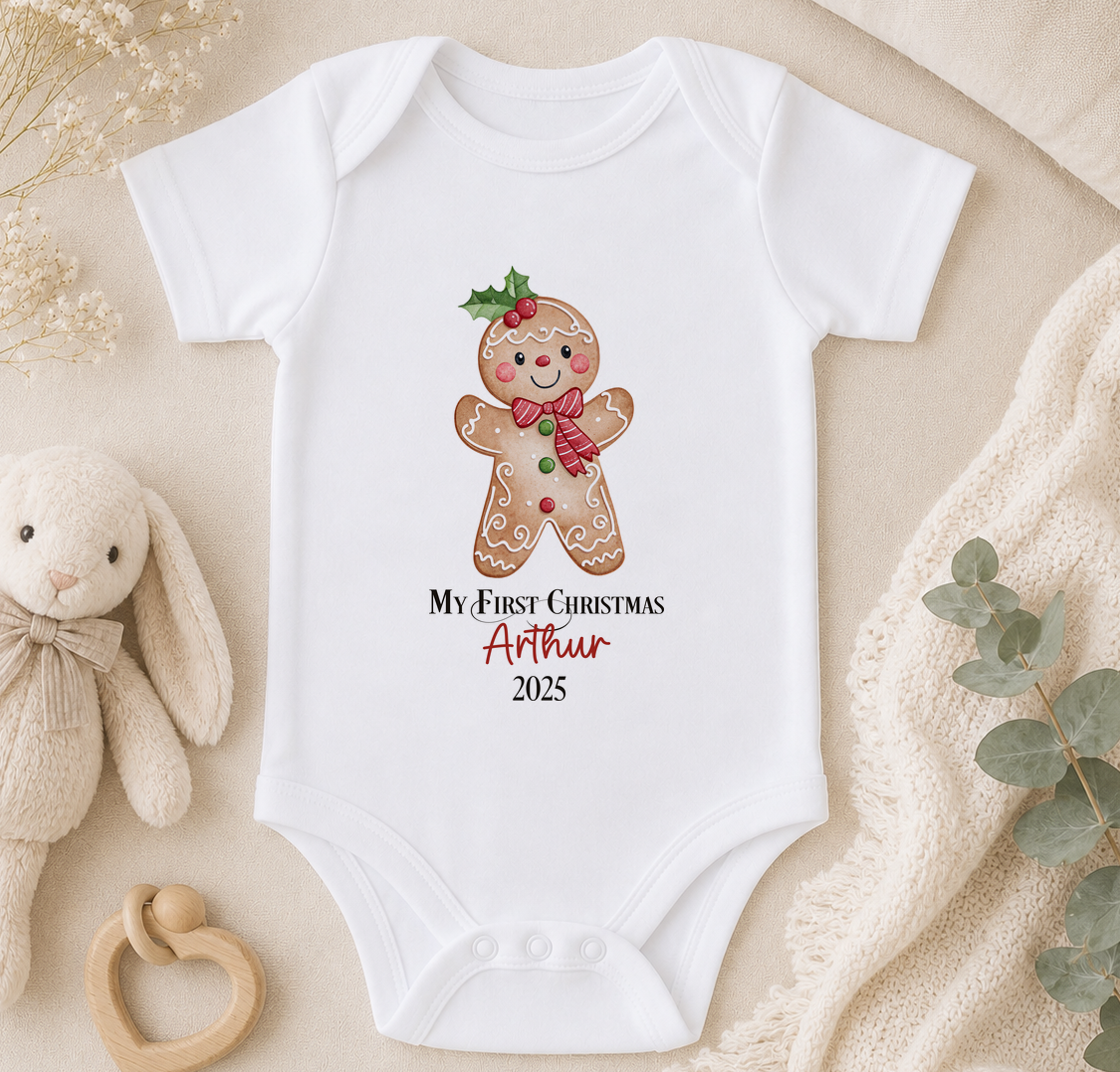 A white onesie with a gingerbread man graphic and text, along with a stuffed animal and some greenery, is displayed on a surface.