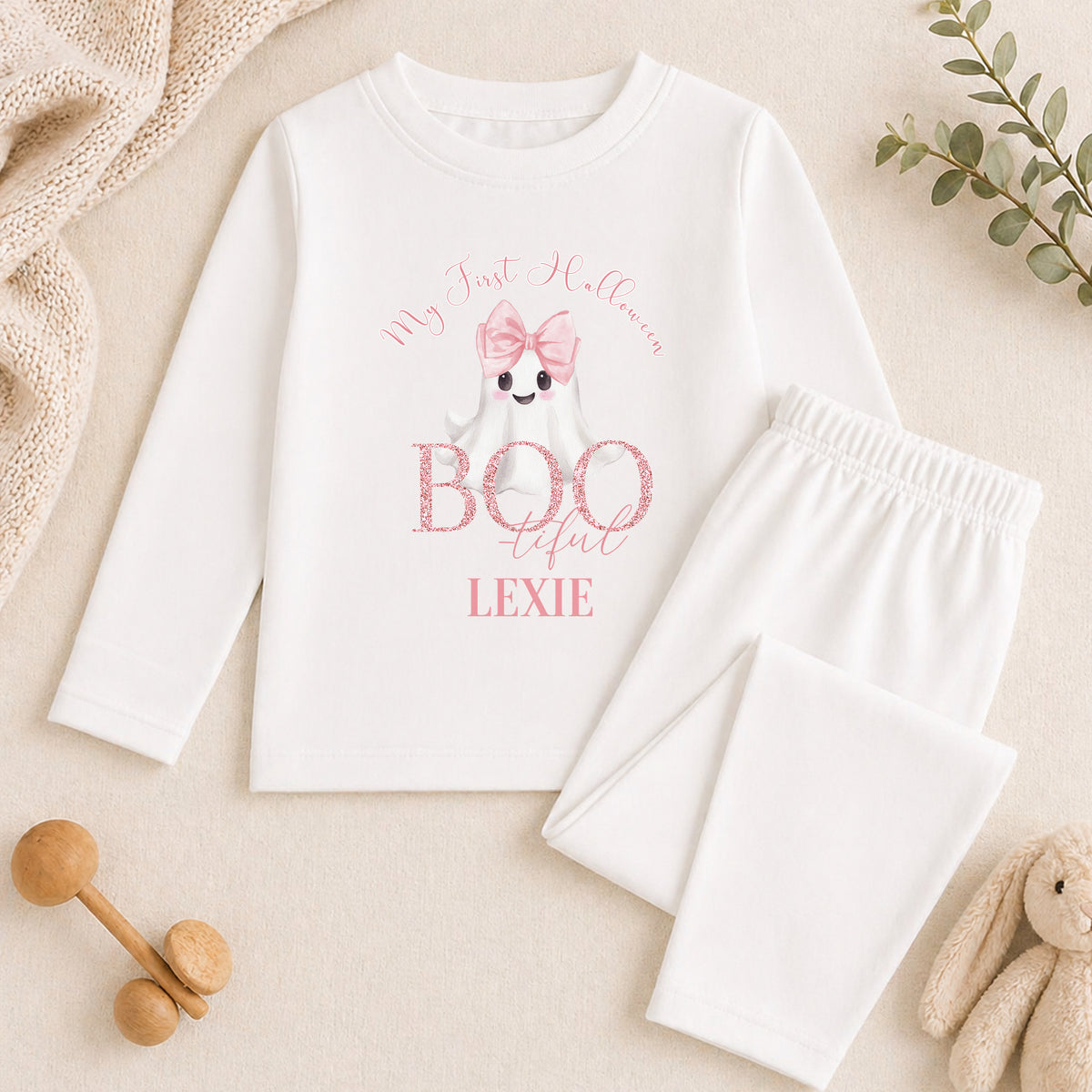 A white long-sleeved shirt with a graphic of a ghost and the text "My first Halloween" printed on it, along with a pink bow. The shirt is accompanied by white pajama pants and a stuffed animal.