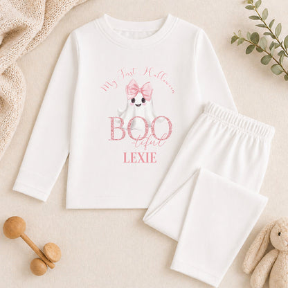 A white long-sleeved shirt with a graphic of a ghost and the text "My first Halloween" printed on it, along with a pink bow. The shirt is accompanied by white pajama pants and a stuffed animal.
