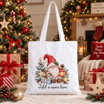 White cotton tote bag printed with a red-hatted Christmas gnome holding a lantern with snowy pine and gold stars, above a name.
