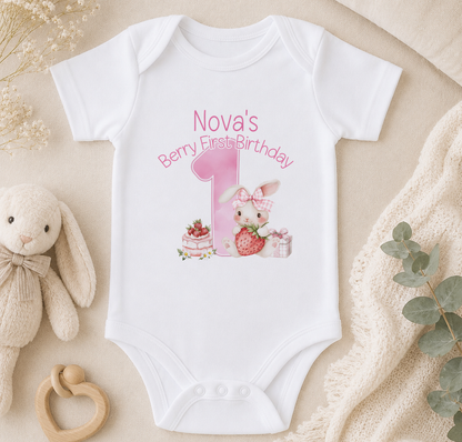 A white onesie with a pink bunny and a pink number 1, along with a berry and a cake, is displayed on a beige surface. There is also a stuffed bunny and some greenery in the background.