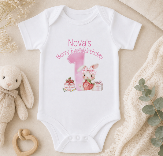 A white onesie with a pink bunny and a pink number 1, along with a berry and a cake, is displayed on a beige surface. There is also a stuffed bunny and some greenery in the background.