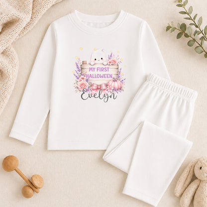 A white long-sleeved shirt with a Halloween-themed graphic and text, along with a pair of white pants, a stuffed animal, and some decorative elements like a plant and a wooden toy.