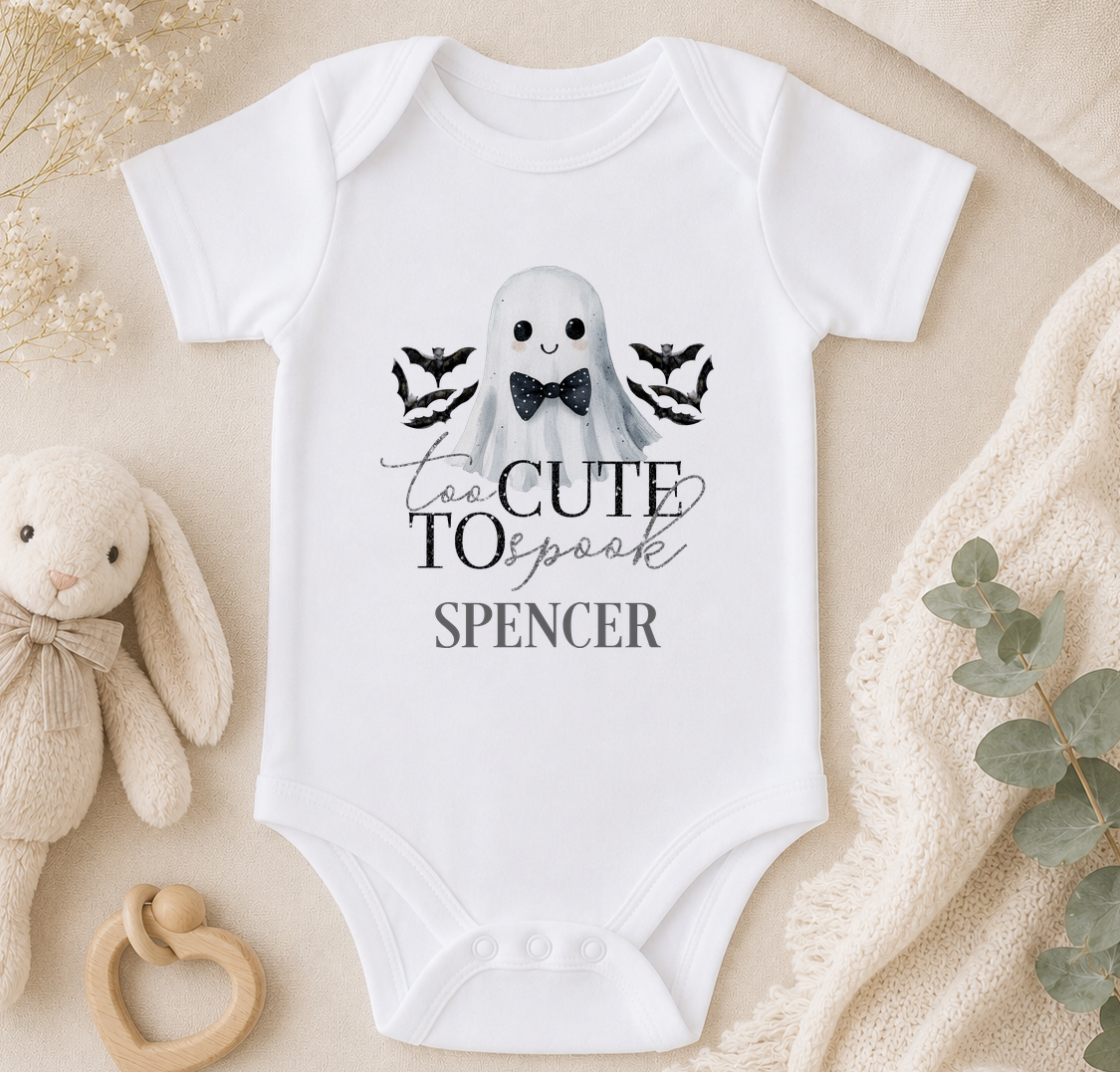 A white baby onesie with a ghost and bats design, along with the text "Too Cute to Spook" and "Spenser" printed on it. The onesie is placed on a beige surface, with a stuffed animal and some greenery nearby.