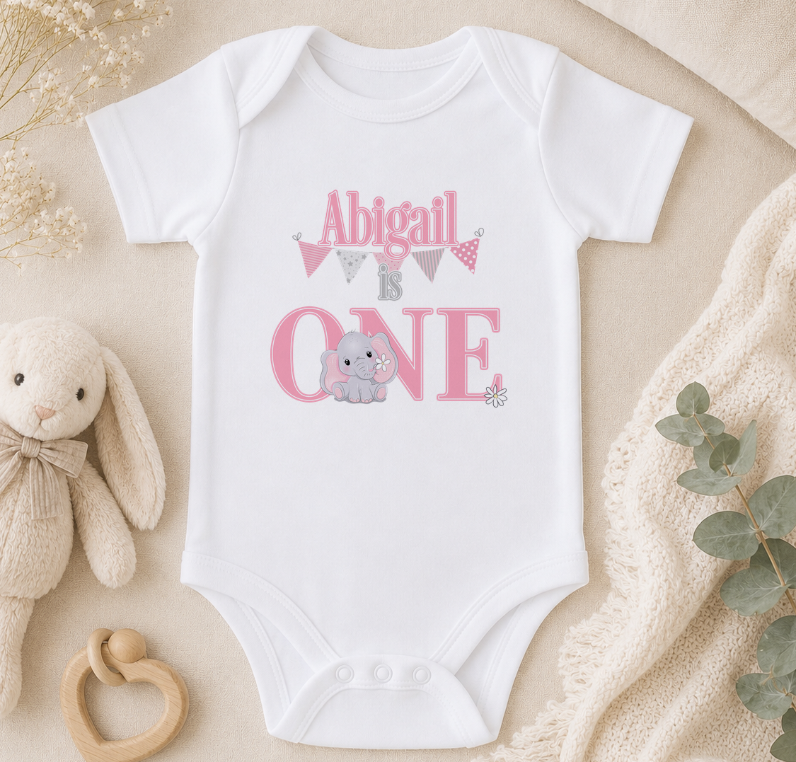 A white onesie with the text "Abigail is one" printed on it, along with an image of an elephant. The onesie is placed on a beige surface, surrounded by a stuffed animal, a baby rattle, and some greenery.