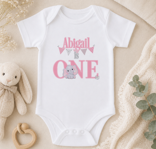 A white onesie with the text "Abigail is one" printed on it, along with an image of an elephant. The onesie is placed on a beige surface, surrounded by a stuffed animal, a baby rattle, and some greenery.