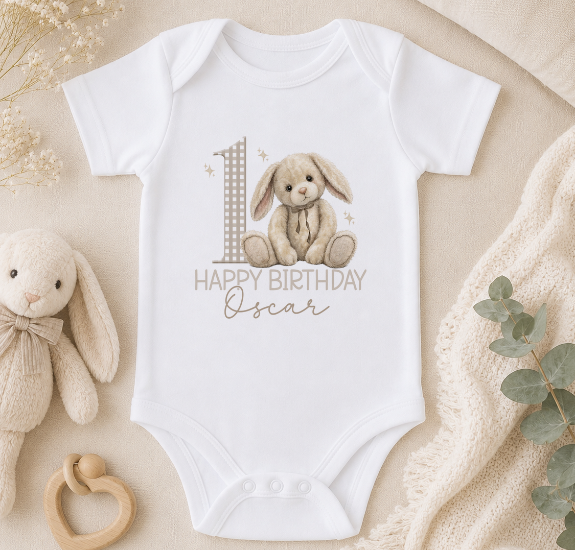 A white onesie with a bunny graphic and the text "Happy Birthday Oscar" printed on it, along with a wooden binky and a small stuffed animal.