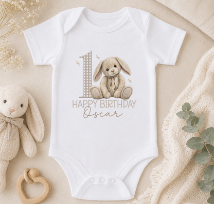 A white onesie with a bunny graphic and the text "Happy Birthday Oscar" printed on it, along with a wooden binky and a small stuffed animal.