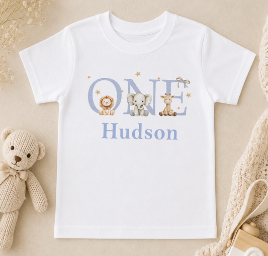 A white t-shirt with the name "Hudson" printed on it, along with illustrations of a lion, elephant, and giraffe. The t-shirt is placed on a beige surface, and there is a stuffed animal and a bottle nearby.