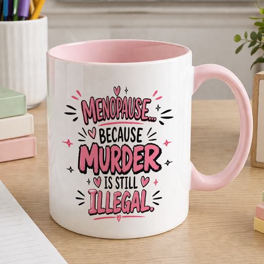 White 11oz ceramic mug with a pink handle and pink inside, printed with 'Menopause... Because Murder is Still Illegal' in pink and black lettering, on a wooden desk.