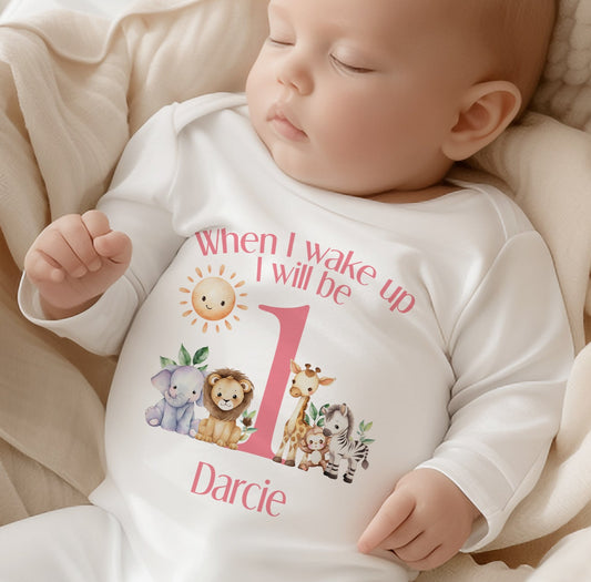 Personalised pink safari sleepsuit and vest set laid out for a first birthday, printed with 'When I Wake Up I Will Be One'.