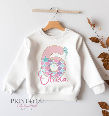 White children's sweatshirt on a hanger printed with a pastel mermaid tail number 6, jellyfish, starfish and the name Olivia