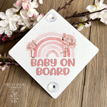Pink flamingo and rainbow Baby on Board car window sign with suction cups displayed on a wooden surface with white blossom