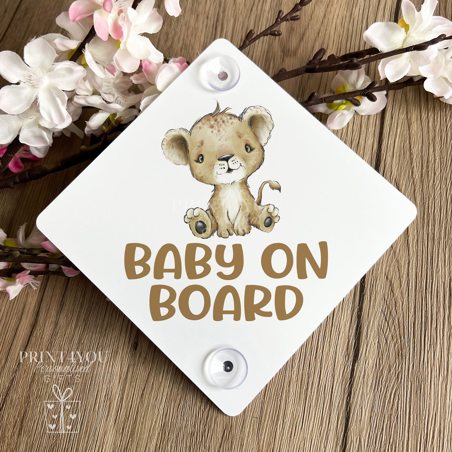 Baby on Board car window sign with baby lion cub design, propped against spring blossom on a wooden surface