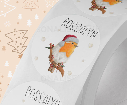 Glossy white round Christmas sticker personalised with a name, printed with a festive robin design, shown on a strip of stickers