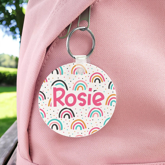 Round colourful rainbow personalised bag tag with the name Rosie, clipped to a pink backpack
