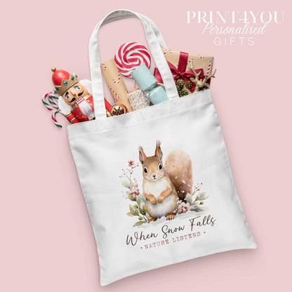 Woodland squirrel Christmas cotton tote bag printed with the words 'When Snow Falls, Nature Listens', shown packed with festive nutcracker and gift wrap props