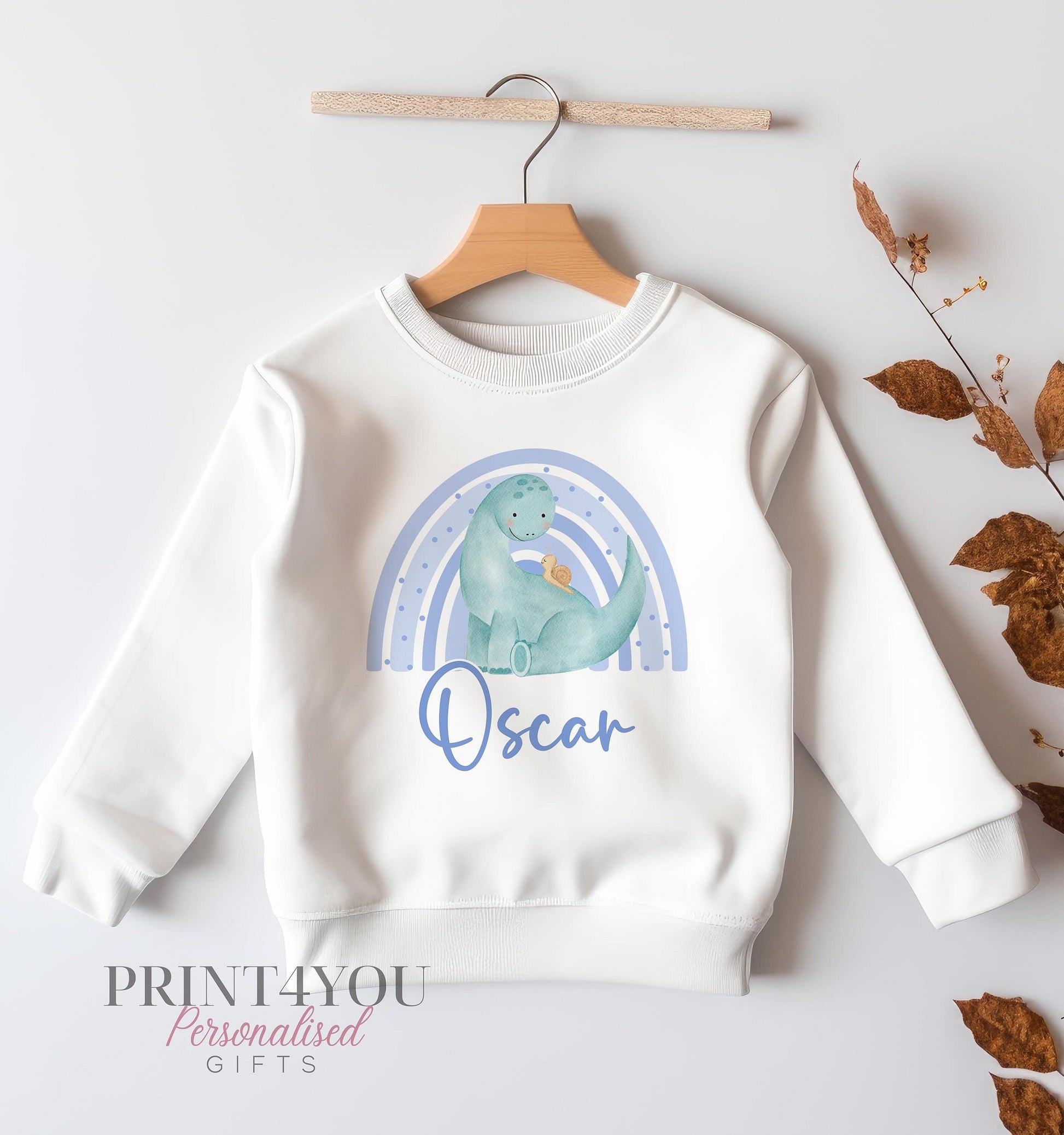 White children's sweatshirt on a hanger printed with a blue watercolour rainbow, a green dinosaur, and the name Oscar in blue script